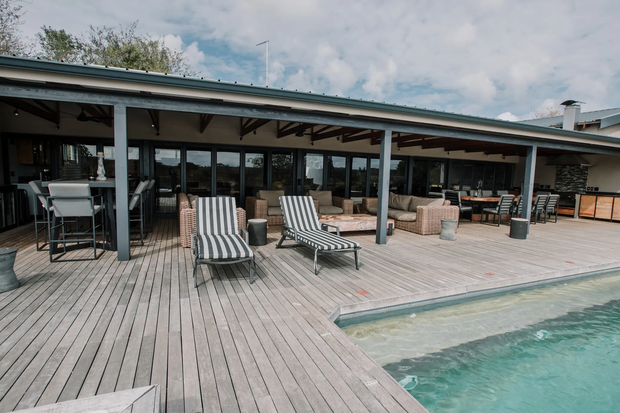 A pool area with a wooden deck, deck chairs and a covered patio with outdoor furniture.